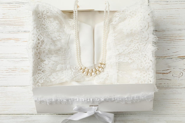 STEP 4: Additional Items—a white lace garment folded in a box topped with a pearl necklace, finished with a white ribbon and placed on a white wooden surface.